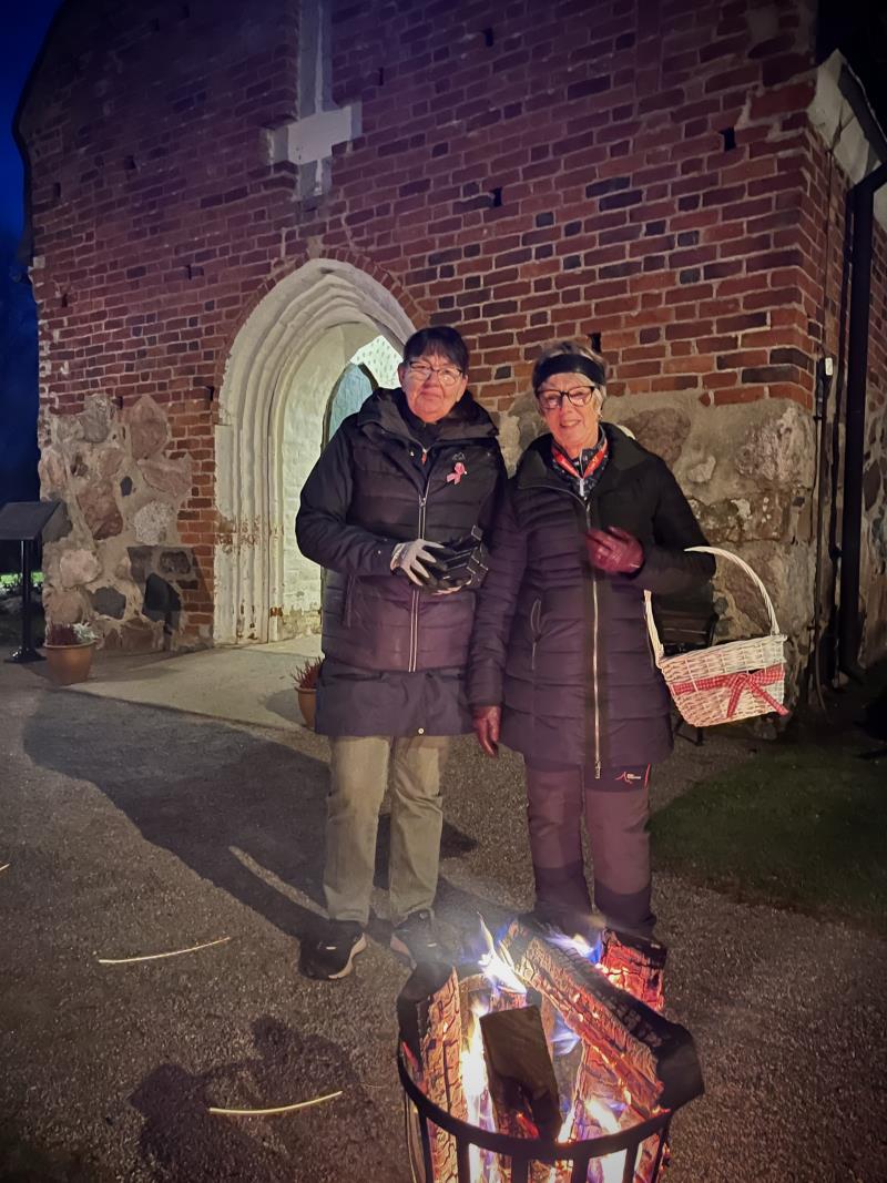 Två kvinnliga volontärer står bredvid varandra utanför Österlövsta kyrka. De de har kronljus i handen som de delar ut till besökare och framför dem brinner en eldkorg.