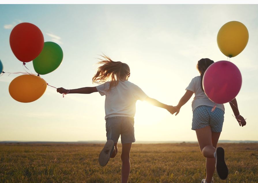 Två barn springer med ballonger i handen mot solen.