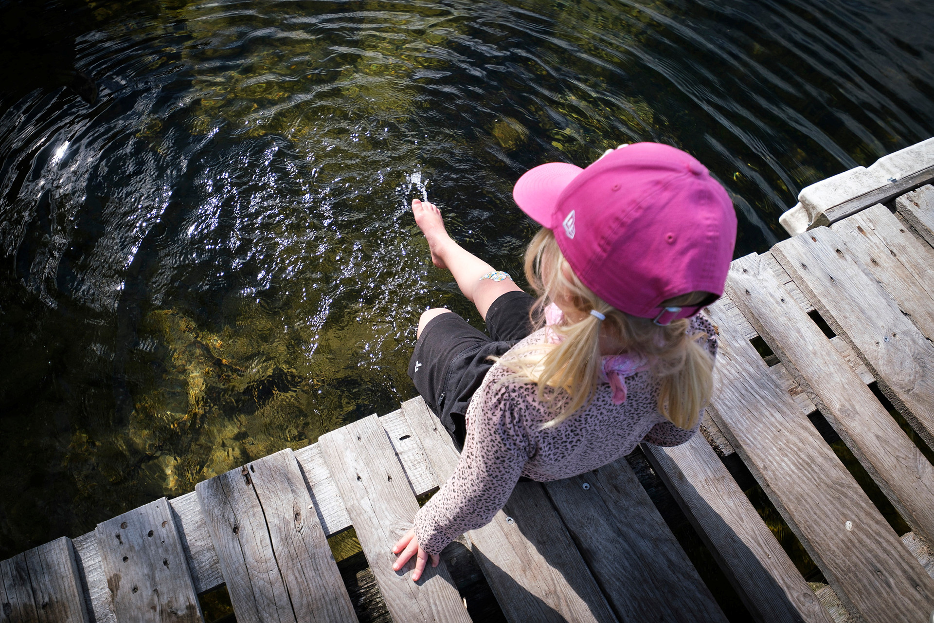 En liten flicka med rosa keps sitter på en brygga och doppar i tårna i vattnet.