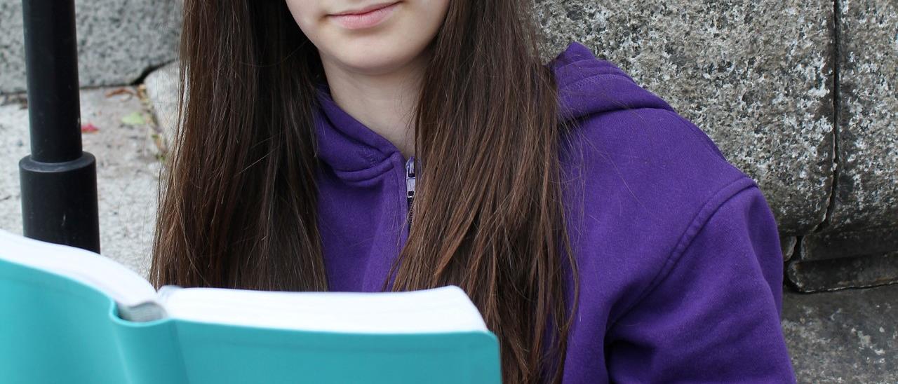En tonårsflicka sitter lutad mot en stenmur och läser ur en turkos bibel.