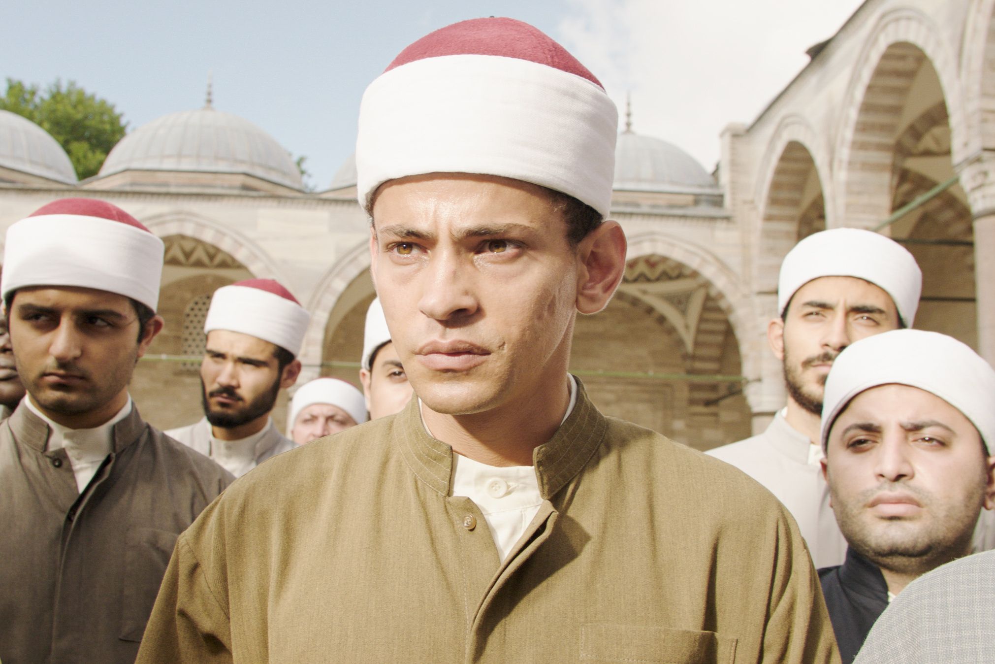 Foto från filmen där huvudpersonen står främst med ett antal män bakom sig, och en moskéliknande byggnad i bakgrunden.