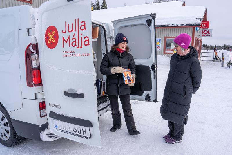 En vit skåpbil står i vinterlandskap och en kvinna delar ut en bok till besökaren.