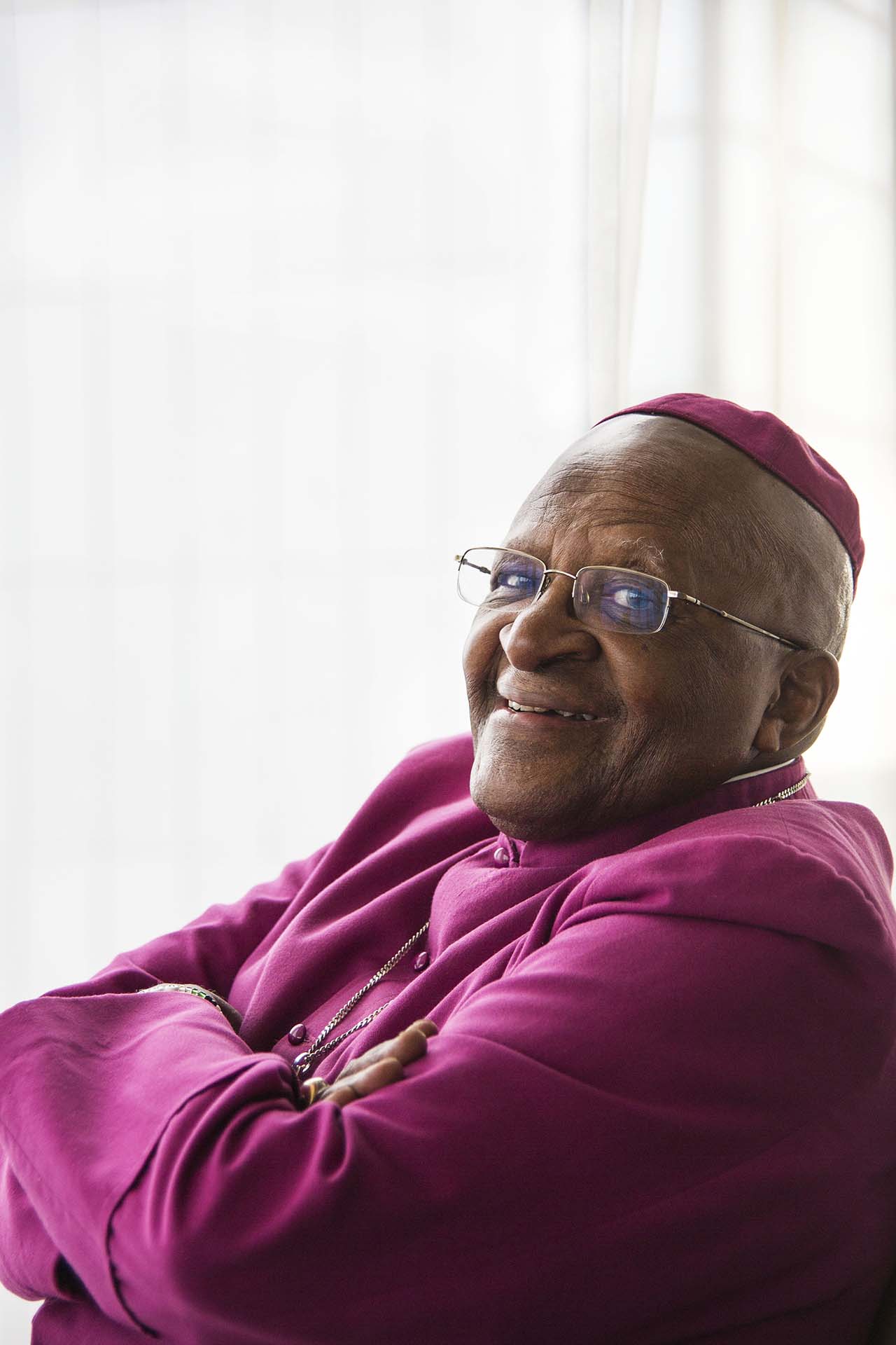 Leende Desmond Tutu i lila skjorta, med armarna i kors.