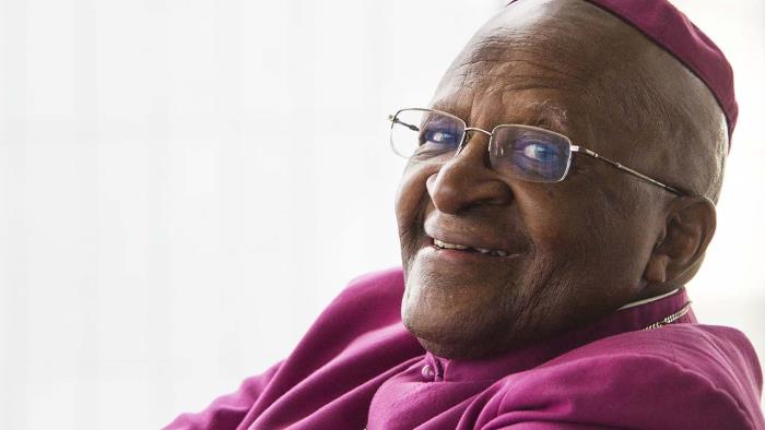 Leende Desmond Tutu i lila skjorta, med armarna i kors.