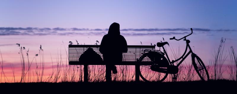 Silhuett av en person sittande på en parksoffa med en cykel bredvid. Himlen är blå och rosa.