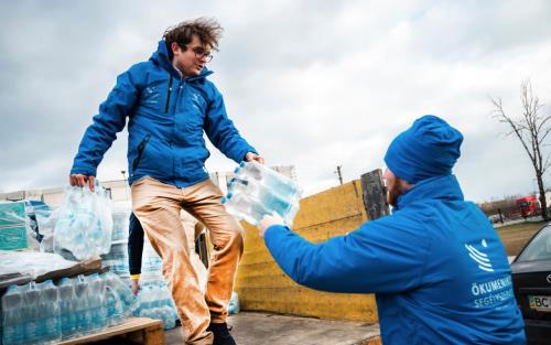 Två män i blå hjälporganisationsjackor lastar av vattenflaskor från ett lastbilsflak.