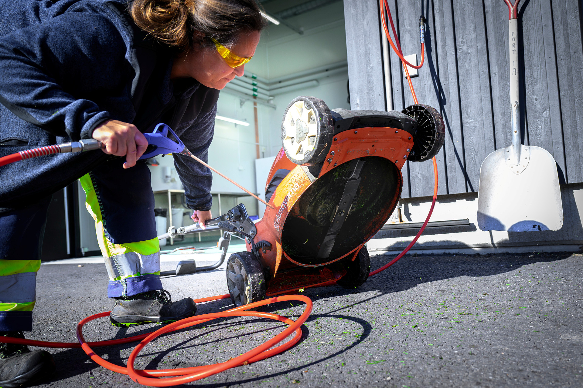 En kyrkogårdsarbetare har vänt upp en elektrisk gräsklippare som hon håller på att rengöra med ett verktyg. Hon har gultonade skyddsglasögon på sig och arbetskläder med reflexdetaljer på.   