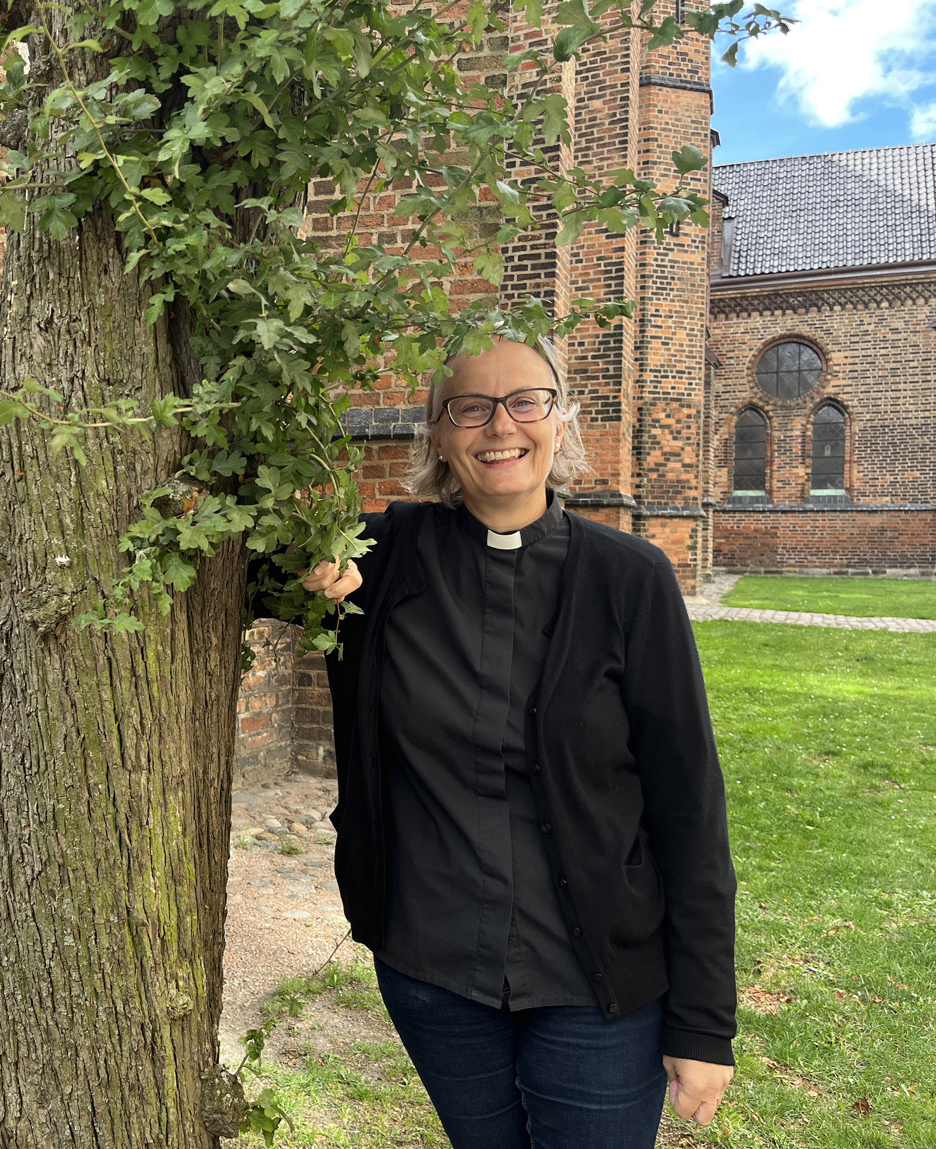 Kvinna med glasögon, svartklädd står vid ett träd och kyrka i bakgrunden.