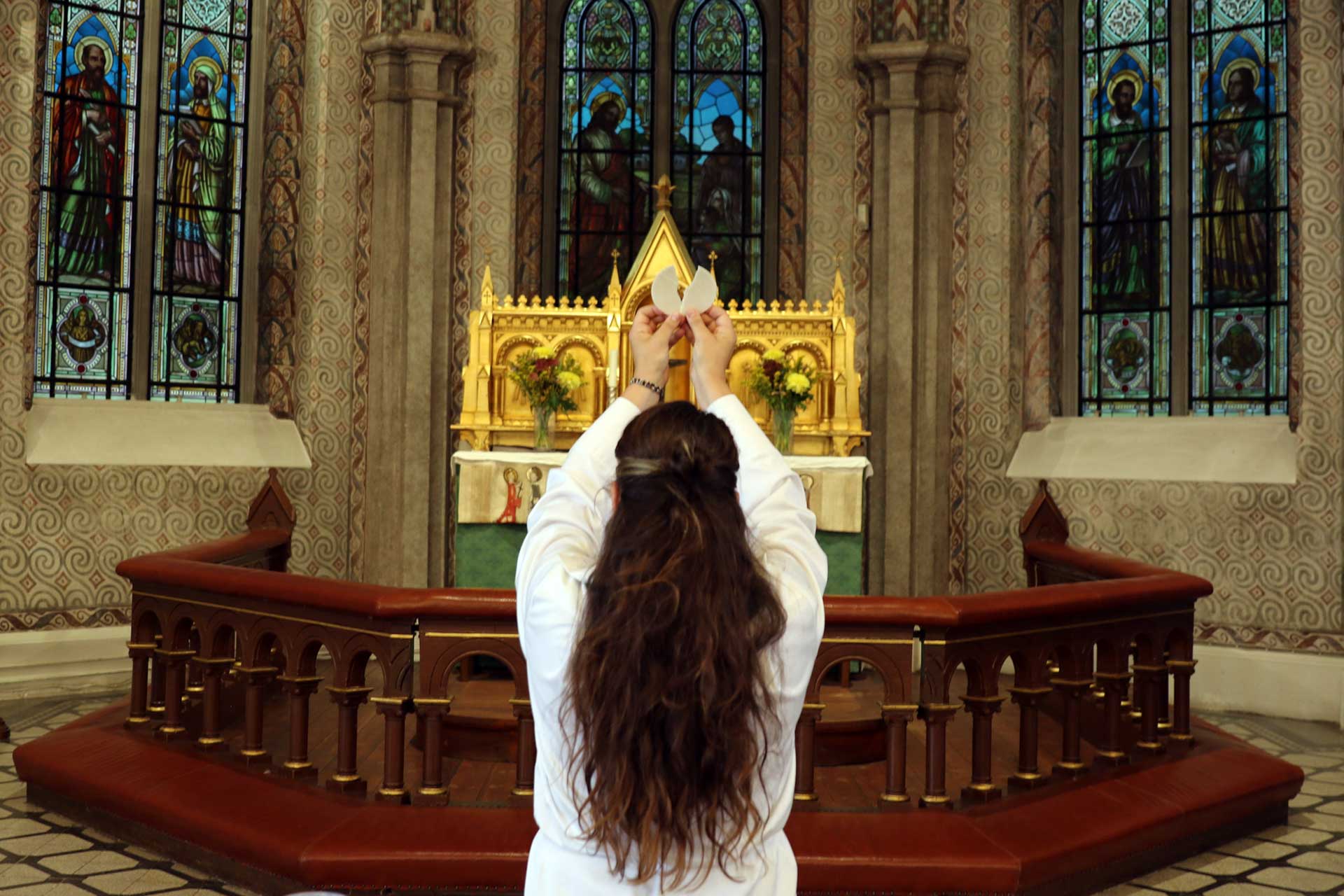 Prästklädd kvinna med långt hår framför guldigt altare och altarring i mörkt trä