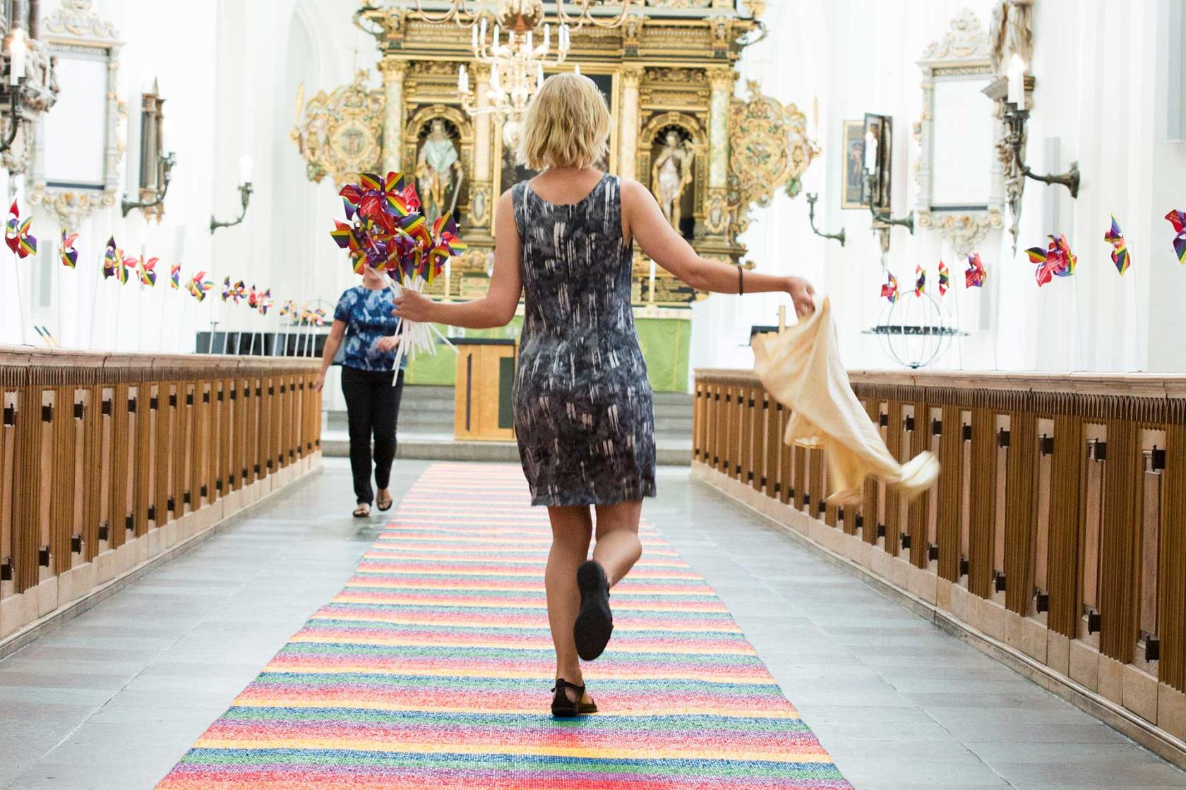 Kvinna går på en regnbågsmatta i mittgången i kyrkan. I handen har hon regnbågssnurror. 