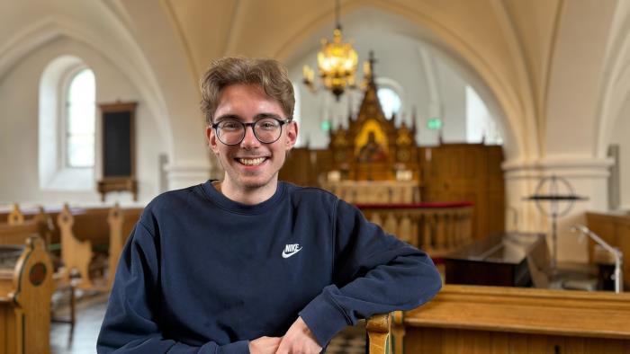 Ung man med glasögon och blå collegetröja med armen lutad mot kyrkbänk.