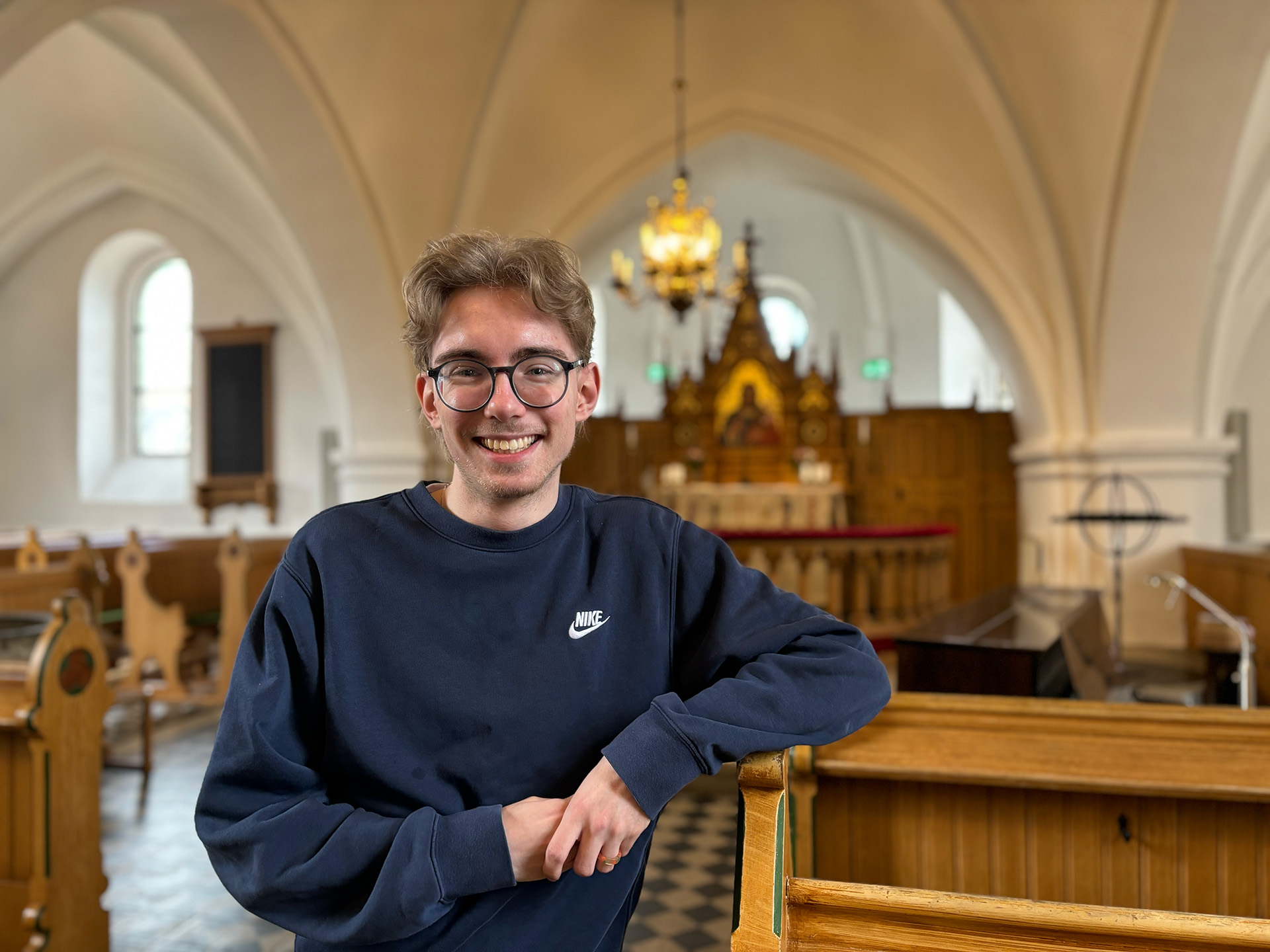 Ung man med glasögon och blå collegetröja med armen lutad mot kyrkbänk.