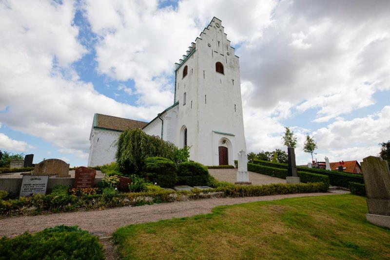 Vitkalkad kyrka med torn med trappstensgavel ligger uppe på en kulle. Runt om finns gravstenar, gräs och grusgångar.