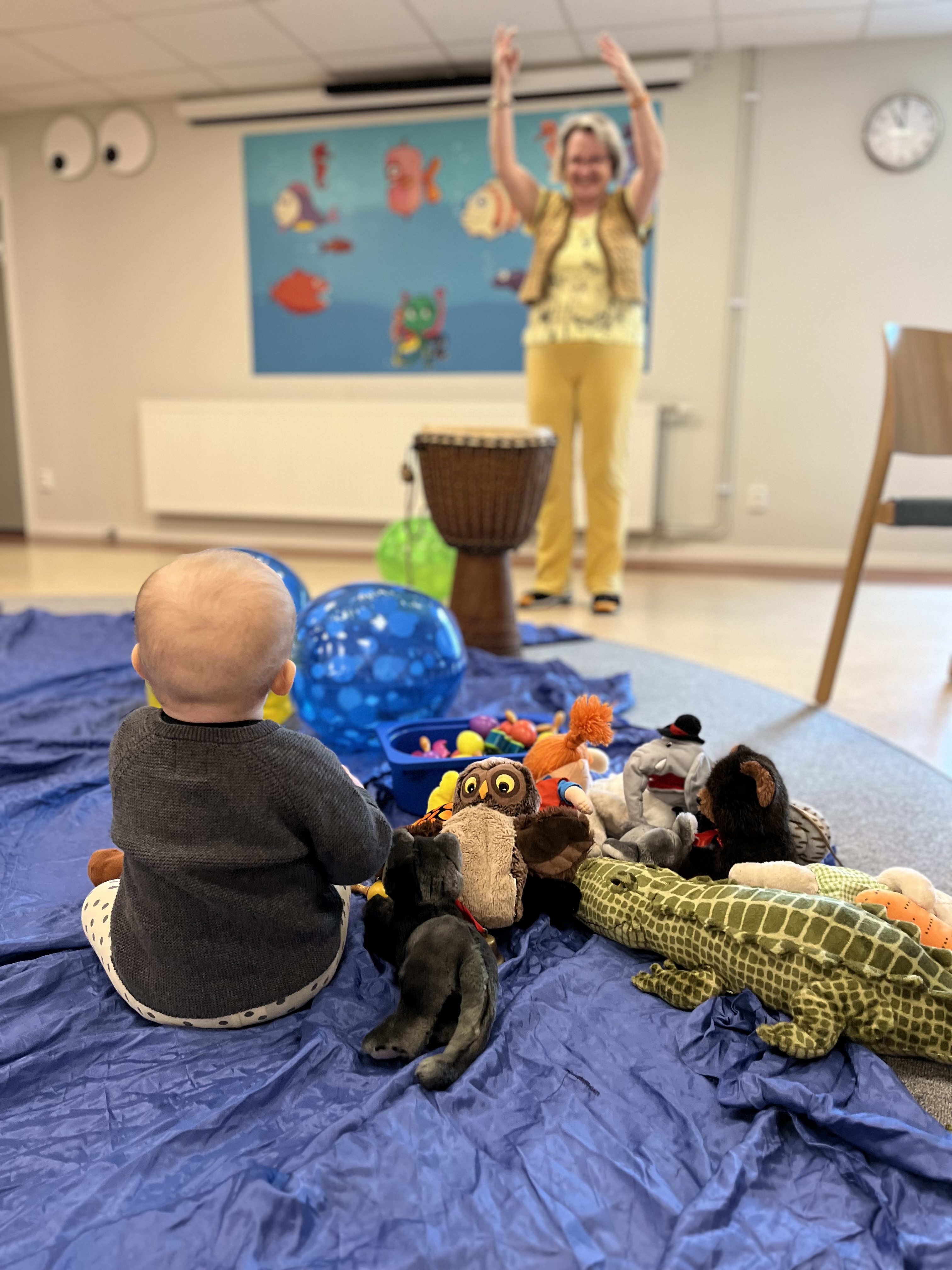 Ett barn sitter på ett stort blått tyg och tittar på många gosedjur och badbollar. Framför barnet står musikern Anna Lena och klappar händer