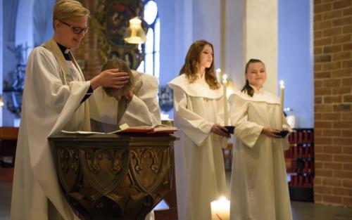 En präst döper en person, två ungdomar står en bit bort och håller i ljus. Alla är klädda i vitt. 