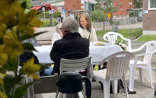 Personer sitter utomhus vid bord och fikar. I förgrunden syns en blomma, i bakgrunden en mataffär.