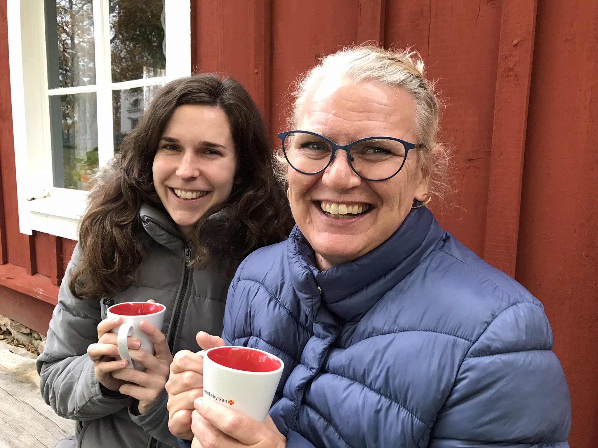 Två kvinnor sitter bredvid varandra med kaffemuggar i händerna. De är utomhus och har vinterjackor på sig.