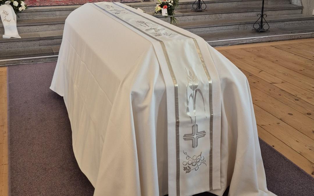 Vitt bårtäcke med broderad löpare draperad över kista i Njurunda kyrka.