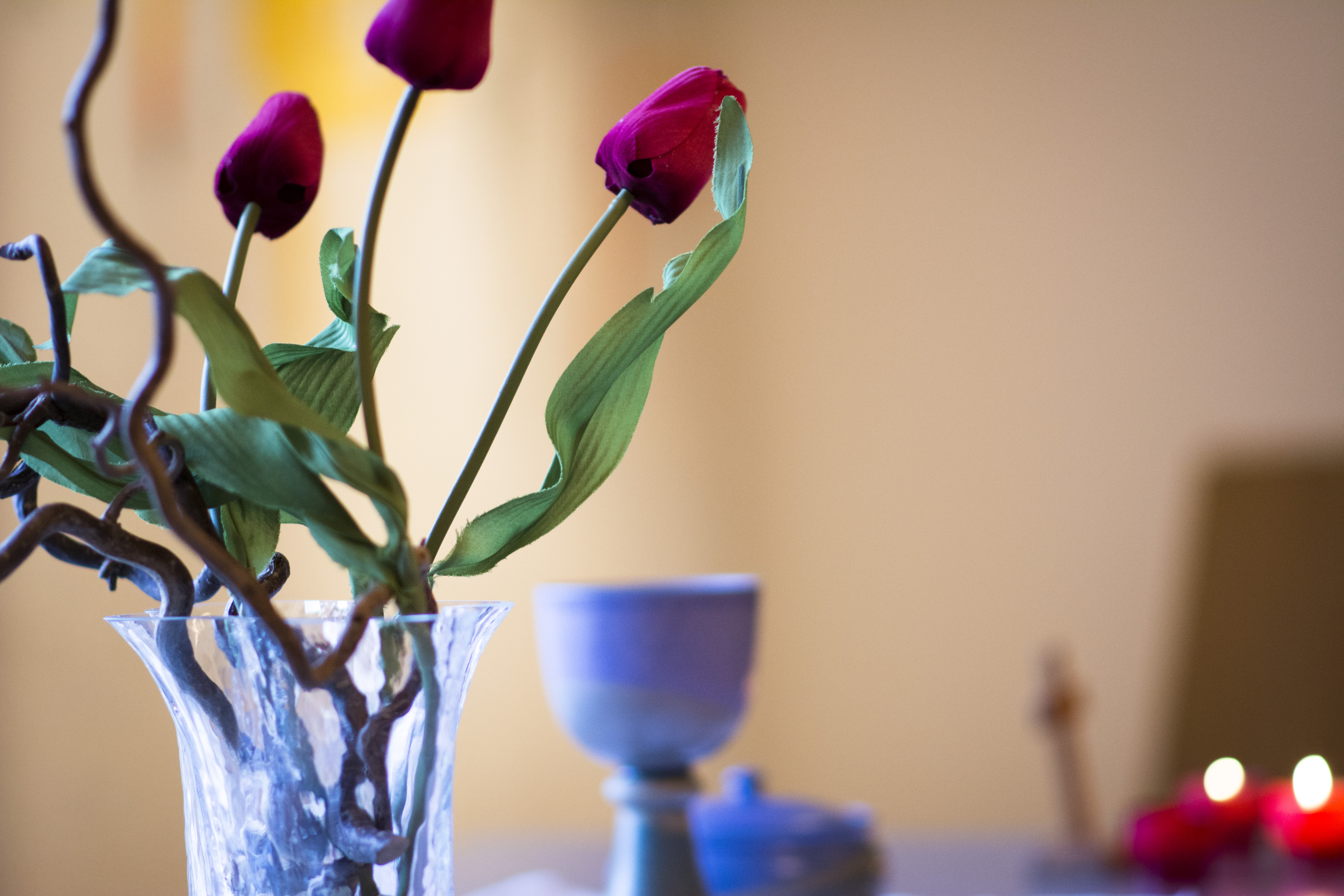 Till vänster syns en vas med röda tulpaner. Vasen är ljusblå och genomskinlig. I bakgrunden till höger syns konturerna  av en kalk, ett träkors och ett par levande taizéljus.
