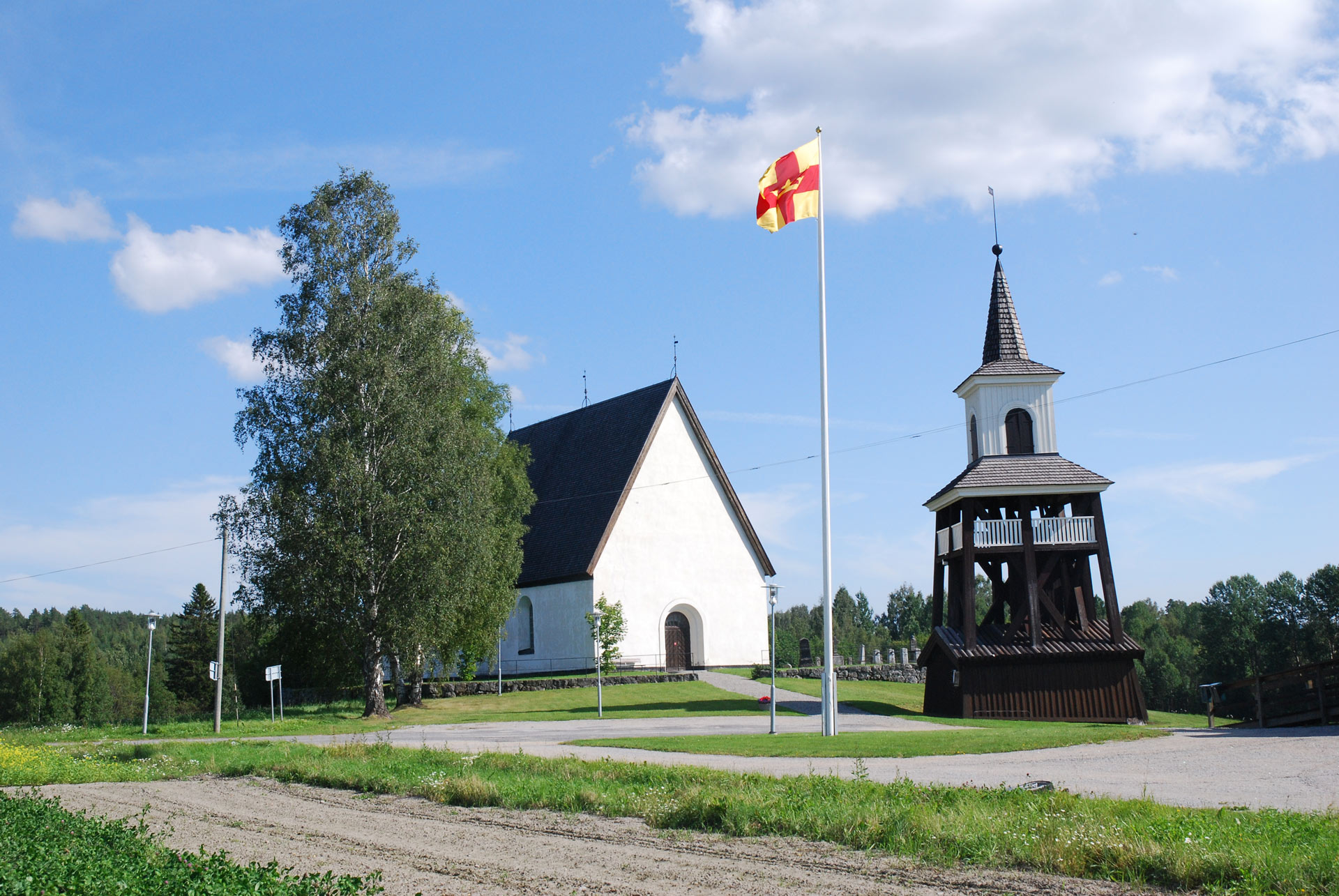 Överlännäs kyrka och klocktorn. Sommarbild. I förgrunden en flaggstång med Svenska kyrkans flagga hissad.