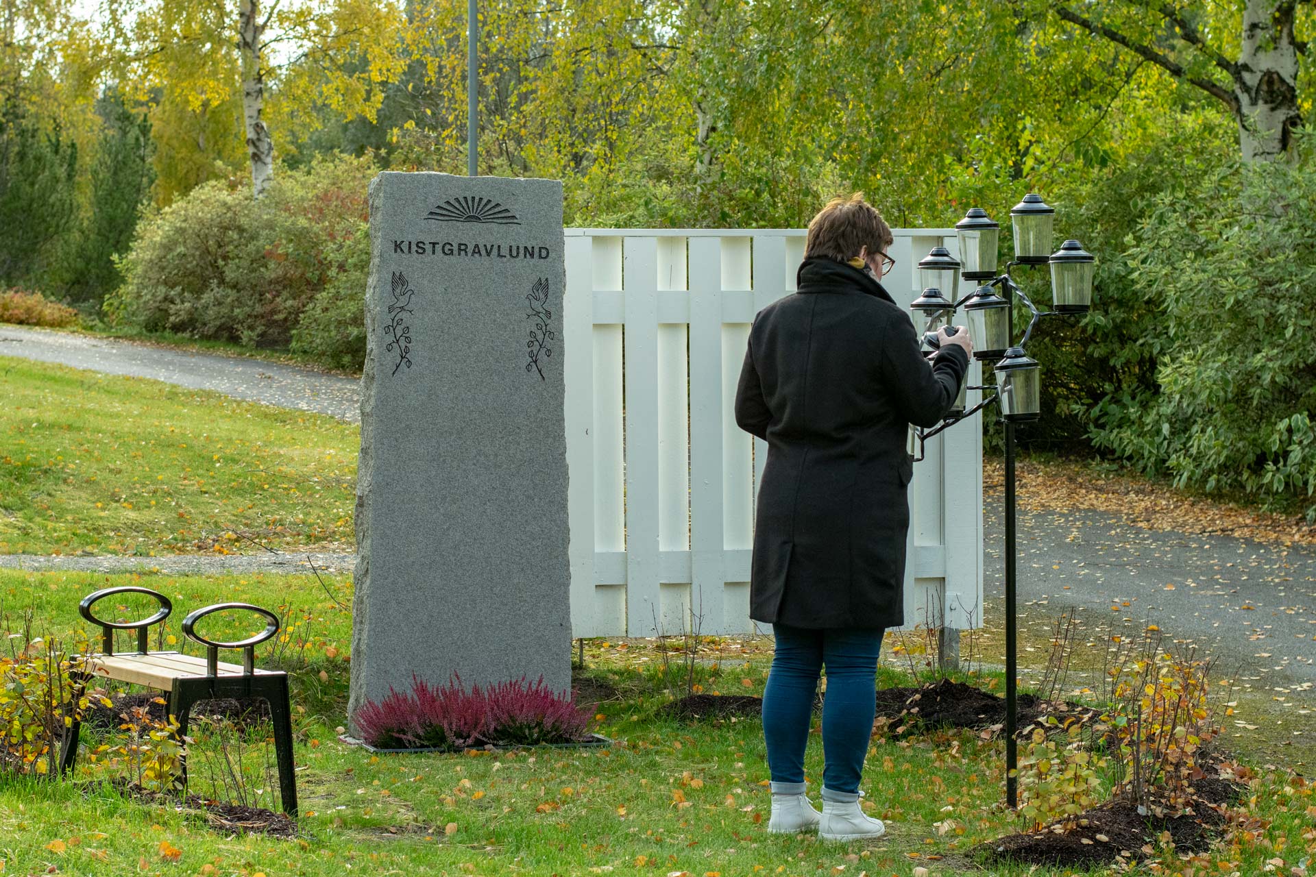 En kvinna står framför en gravsten med texten "Kistgravlund". Hon lyfter på locket till en av ljuslyktorna i en ställning med flera lyktor.  Det är höst.