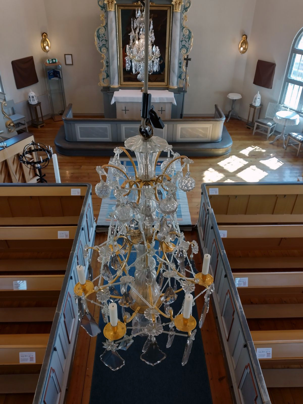 En kristallkrona i rokokostil hänger i en kyrka. Fotot är taget från orgelläktare . Vi ser kronan snett ovanifrån och i bakgrunden altaret och ytterligare en ljuskrona.
