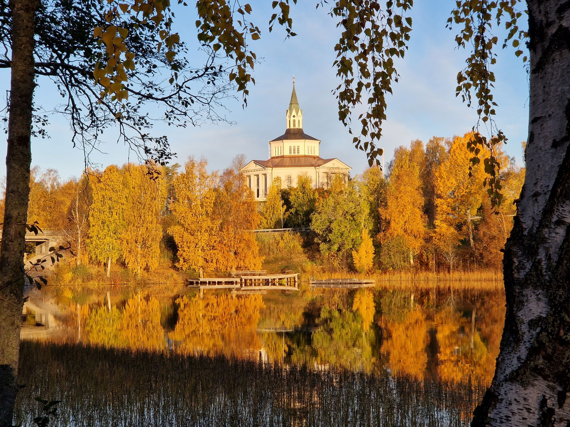 Bild på Själevads kyrka på höjden. I förgrunden en spegelblank Moälv och träden är gyllene gula. 