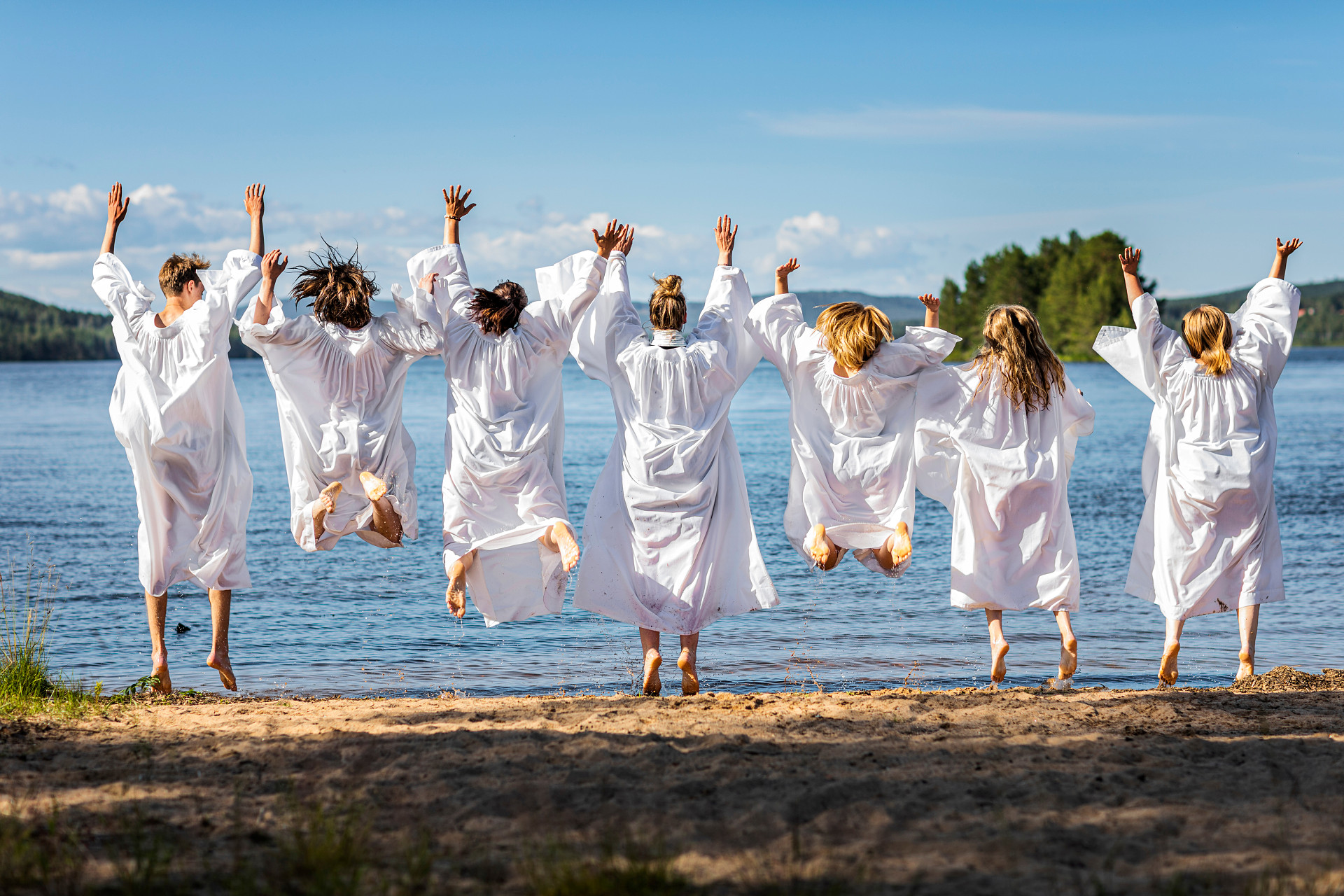 Ungdomar vid en sjö, som tillsammans gör ett hopp, klädda i vita konfirmandkåpor 