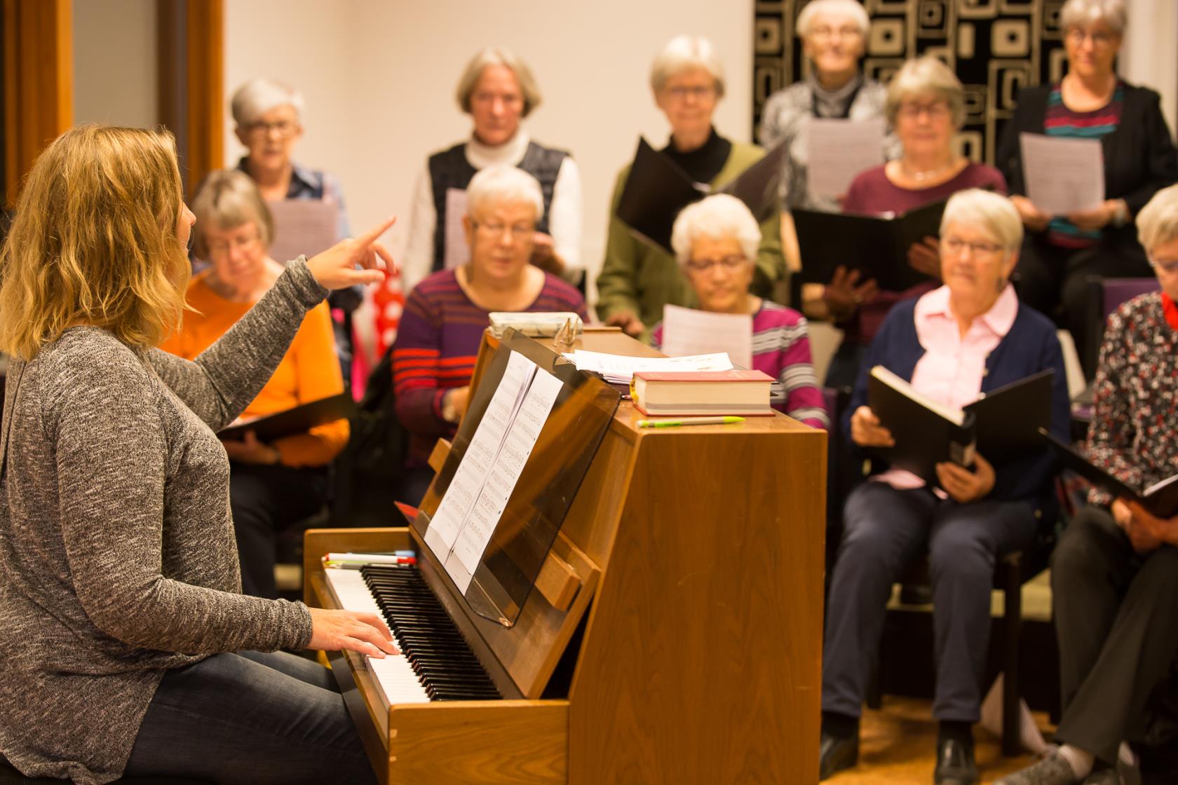 En körledare som sitter vid ett piano och som leder en sångkör.