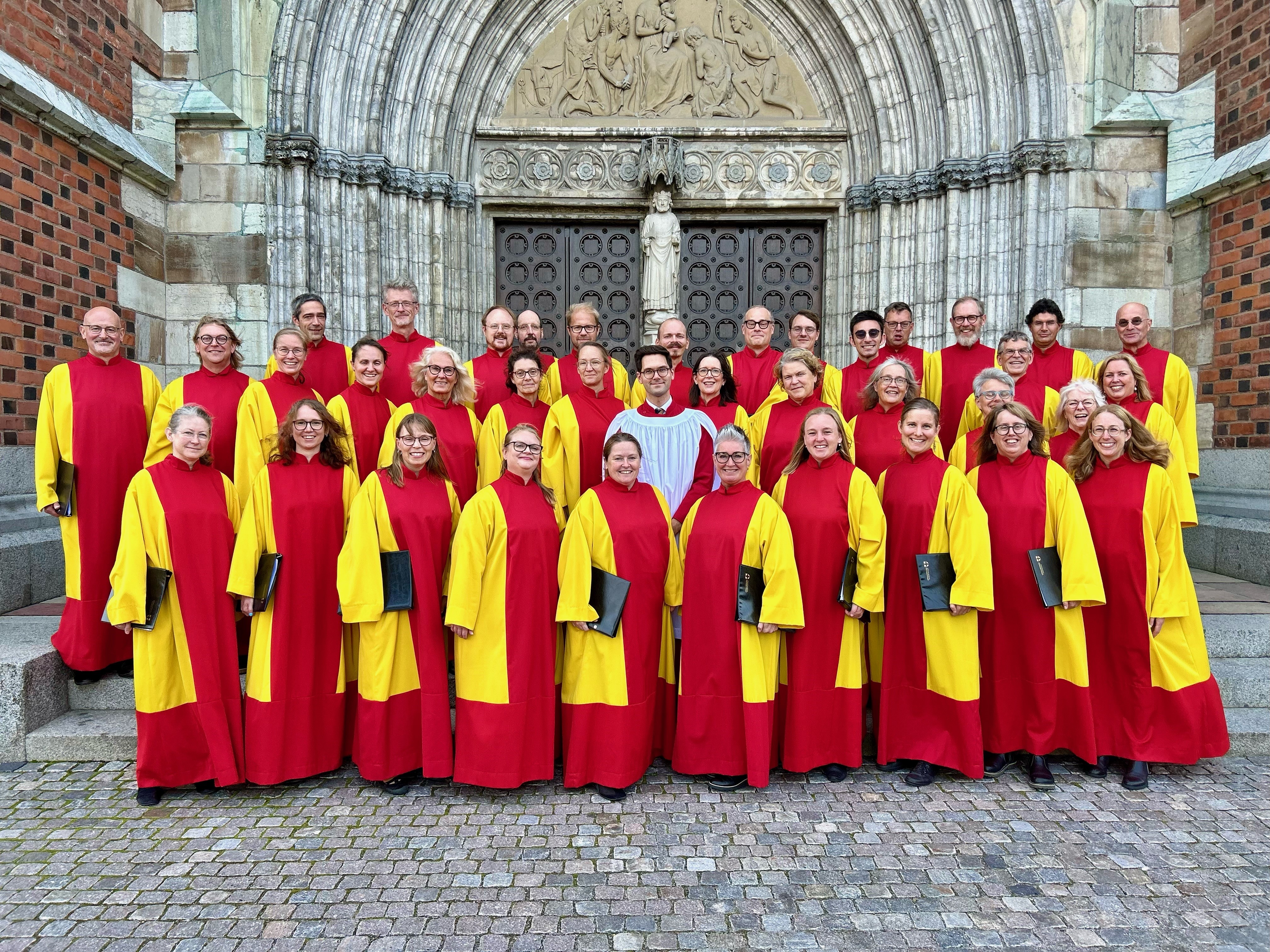 Kören Collegium Cantorum i gula och röda körkåpor står grupperade kring sin körledare Robert Bennesh på trappan utanför Uppsala domkyrkas norra port.