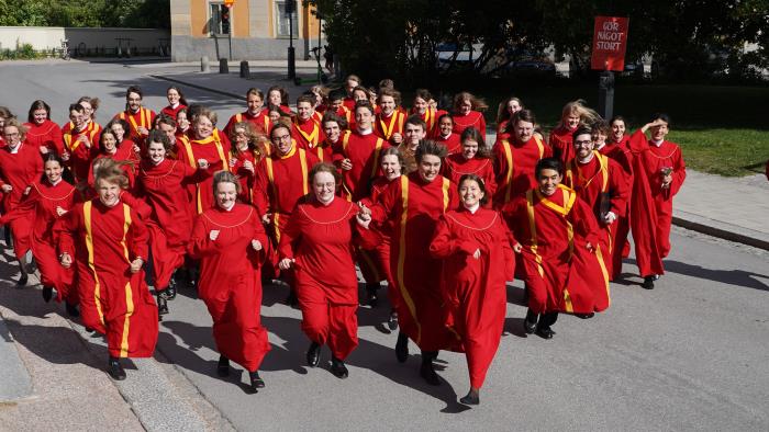 Uppsala domkyrkas ungdomskör springer glatt mot fotografen tvärs över gatan i sommarsolen, iförda röda körkåpor.