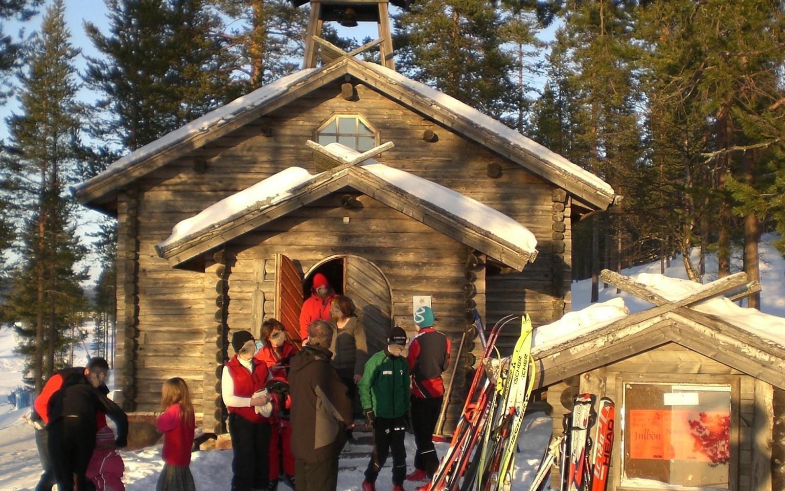 Träkapell i fjällmiljö med människor i skidkläder och skidor utanför.