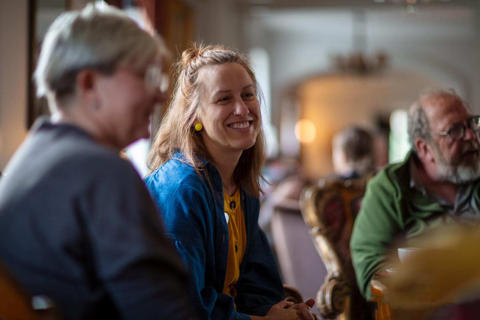 Flera personer i cafémiljö samtalar. Leende kvinna i fokus. 