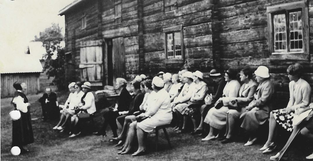 Svartvitt foto på ett tjugotal kvinnor som sitter uppradade utanför ett gammalt trähus