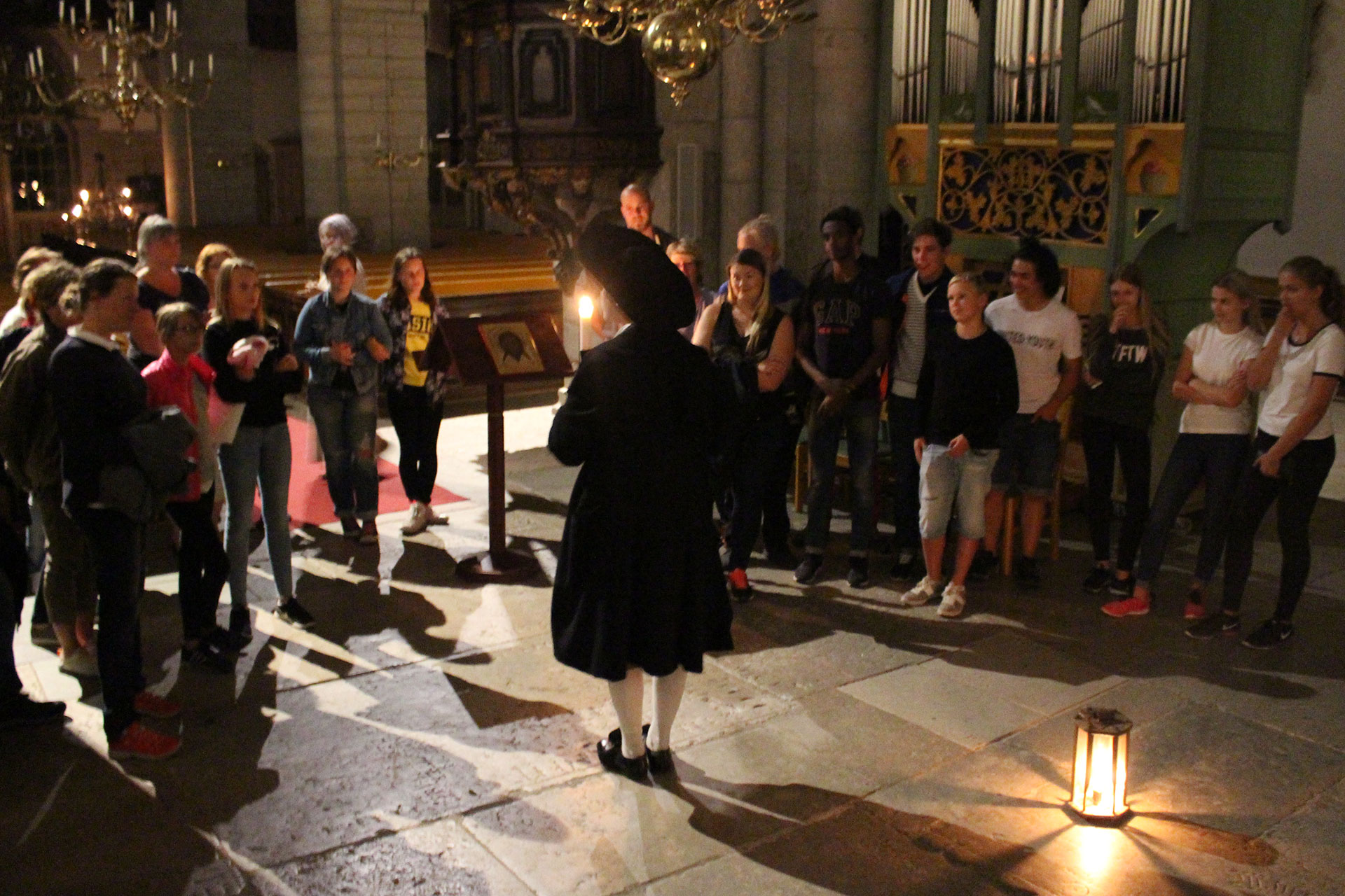 Besökare står i en halvcirkel mitt i kyrkan runt en 1700-talsguide i stor hatt. 