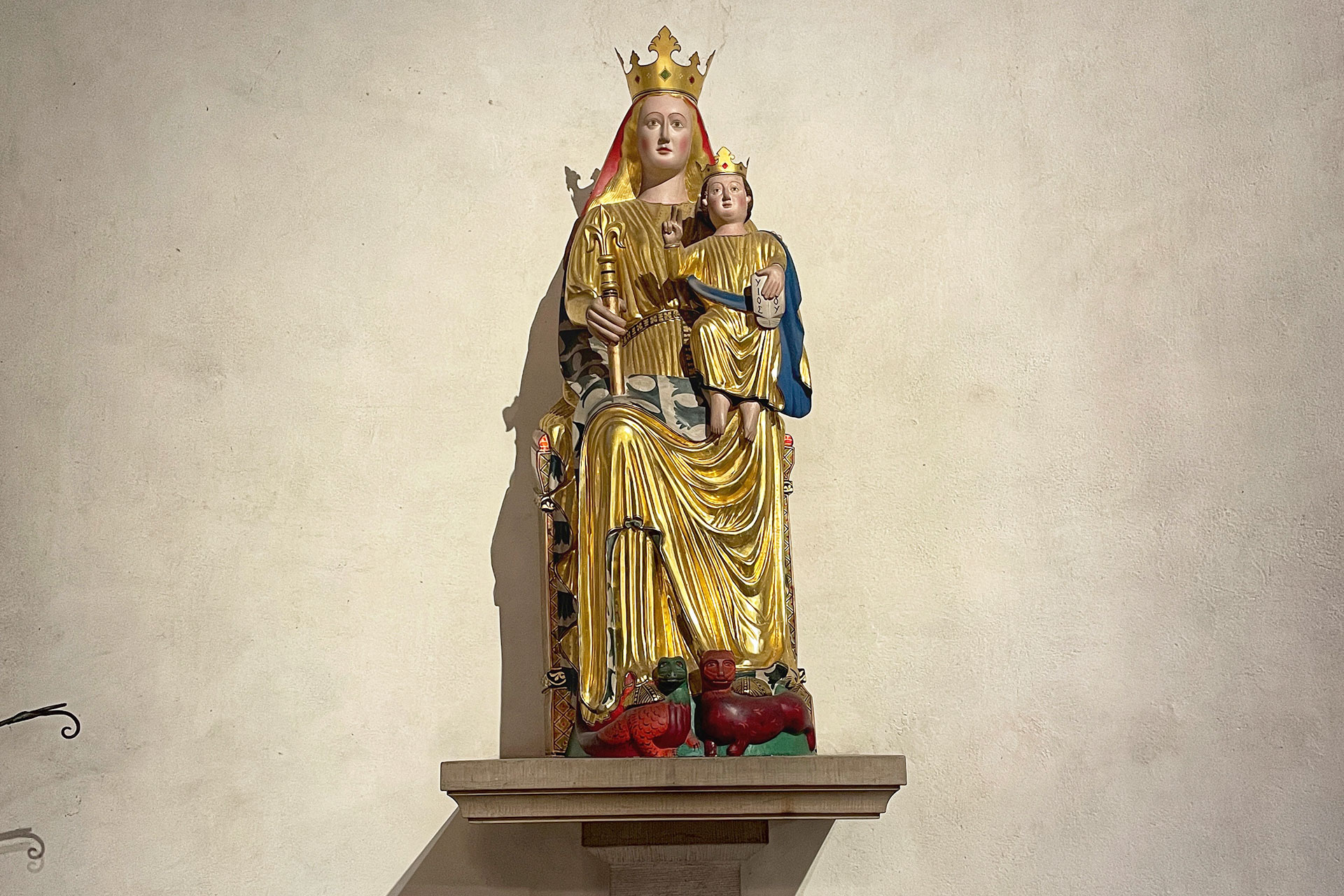 Maria sitter på en tron iförd en gyllene klänning med guldmönstrat smalt bälte och pälsfodrad gyllene mantel med rödbrun krage. I höger hand håller hon en spira. På sitt vänstra knä håller hon Jesubarnet som är klätt i en fotsid, gyllene skjorta med mantel i fodrad blått. 