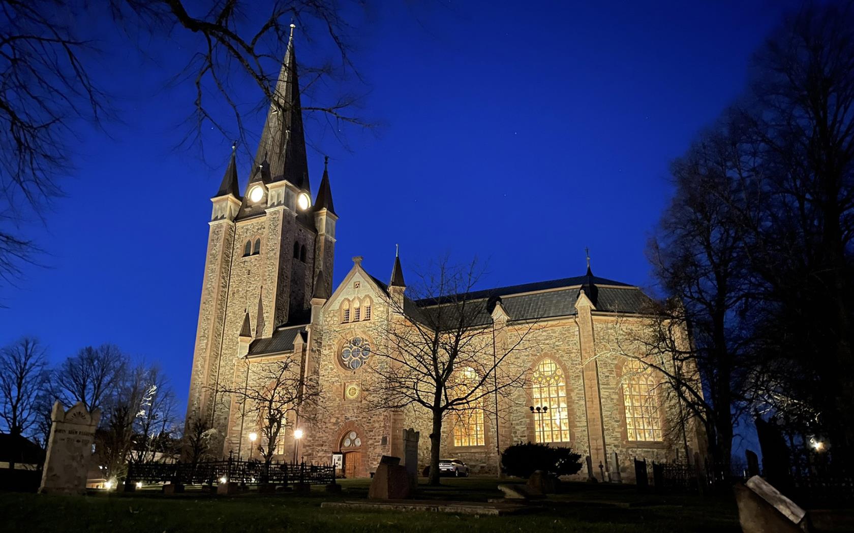 Upplyst stor stenkyrka i rödaktig sten mot en mörkblå natthimmel.