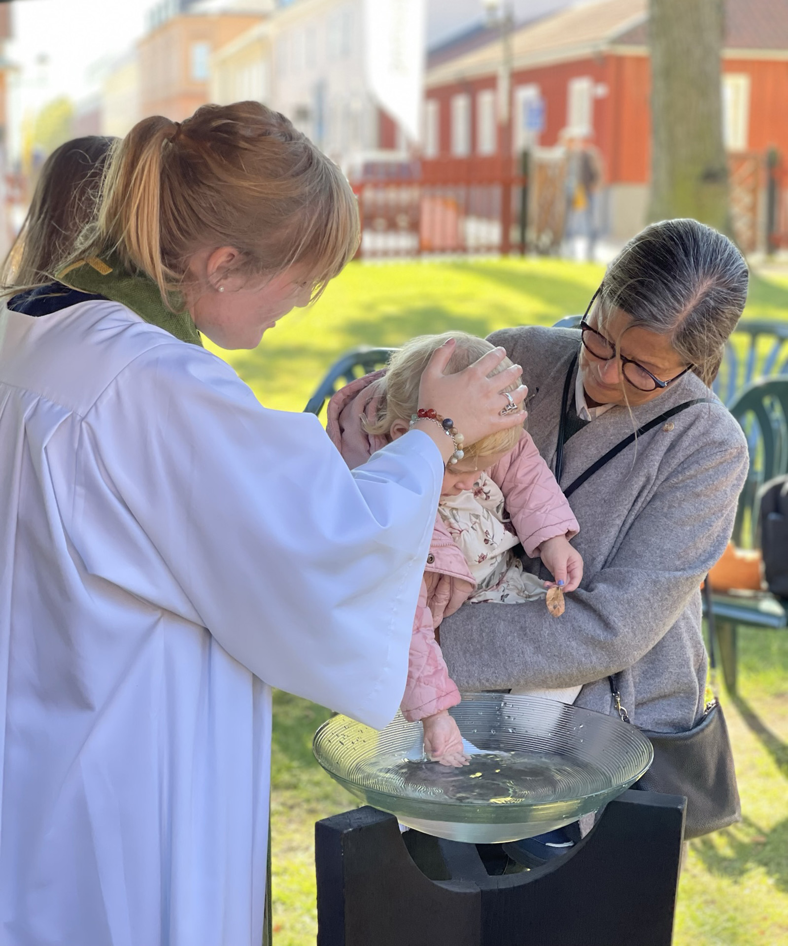 Kvinnlig präst häller vatten på ett barn huvud. Barnet hålls i famnen av sin mormor. Detta är ett utomhusdop.