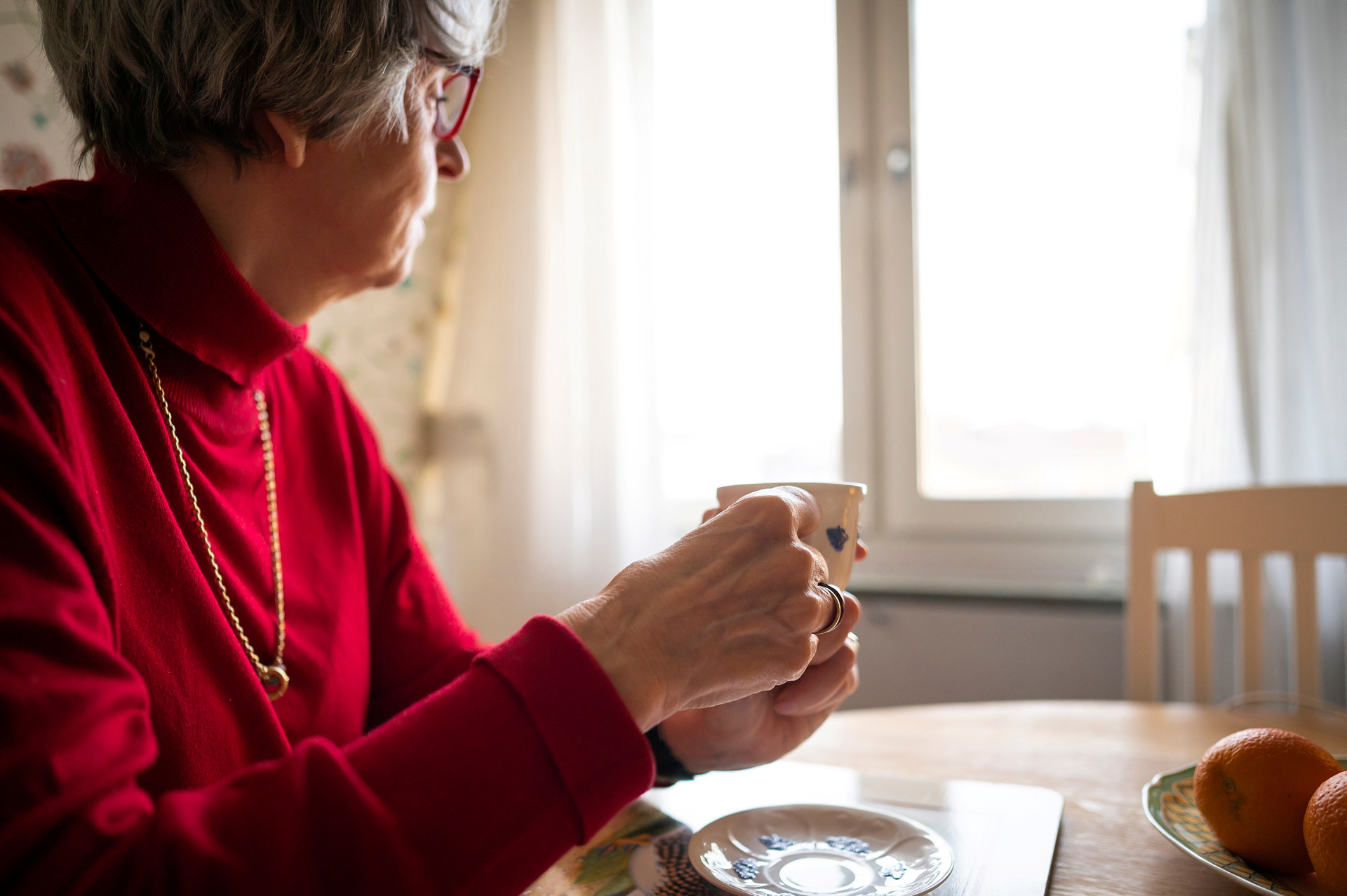 Kvinna i röd tröja dricker en kopp kaffe själv vid köksbordet och tittar ut genom fönstret.