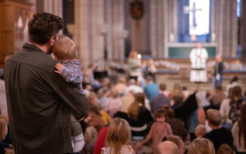 Barn och vuxna i en kyrka, fokus på en man och ett barn som står med ryggen mot kameran och kollar mot prästen