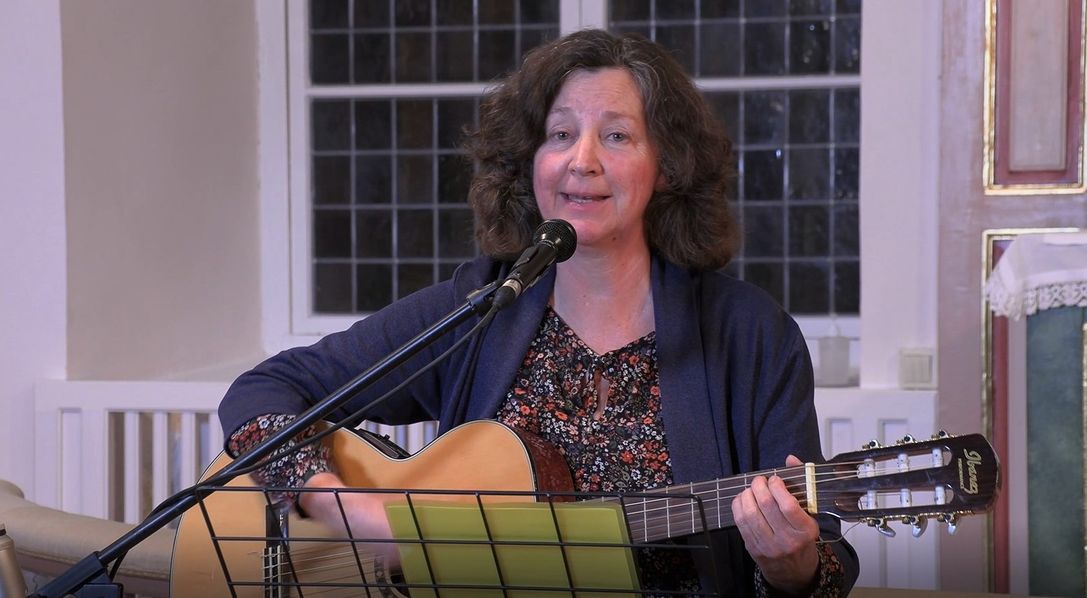 Maria Samuelsson sjunger sitter på en hög pall och sjunger sin Corona-sång samtidigt som hon spelar gitarr. 