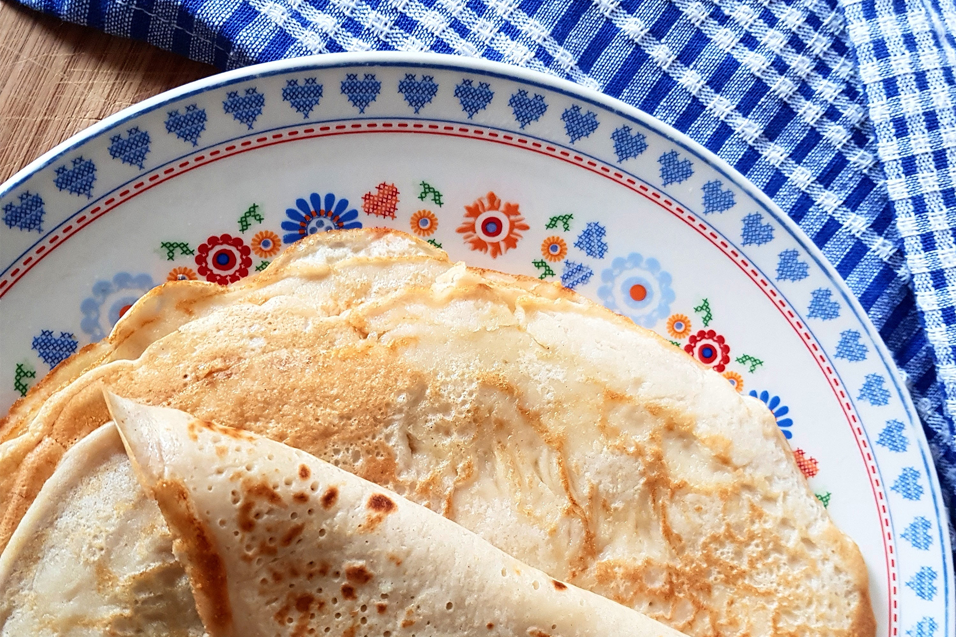 Pannkakor i hög på en blommig porslinstallrik på bord med blåvitrutig duk