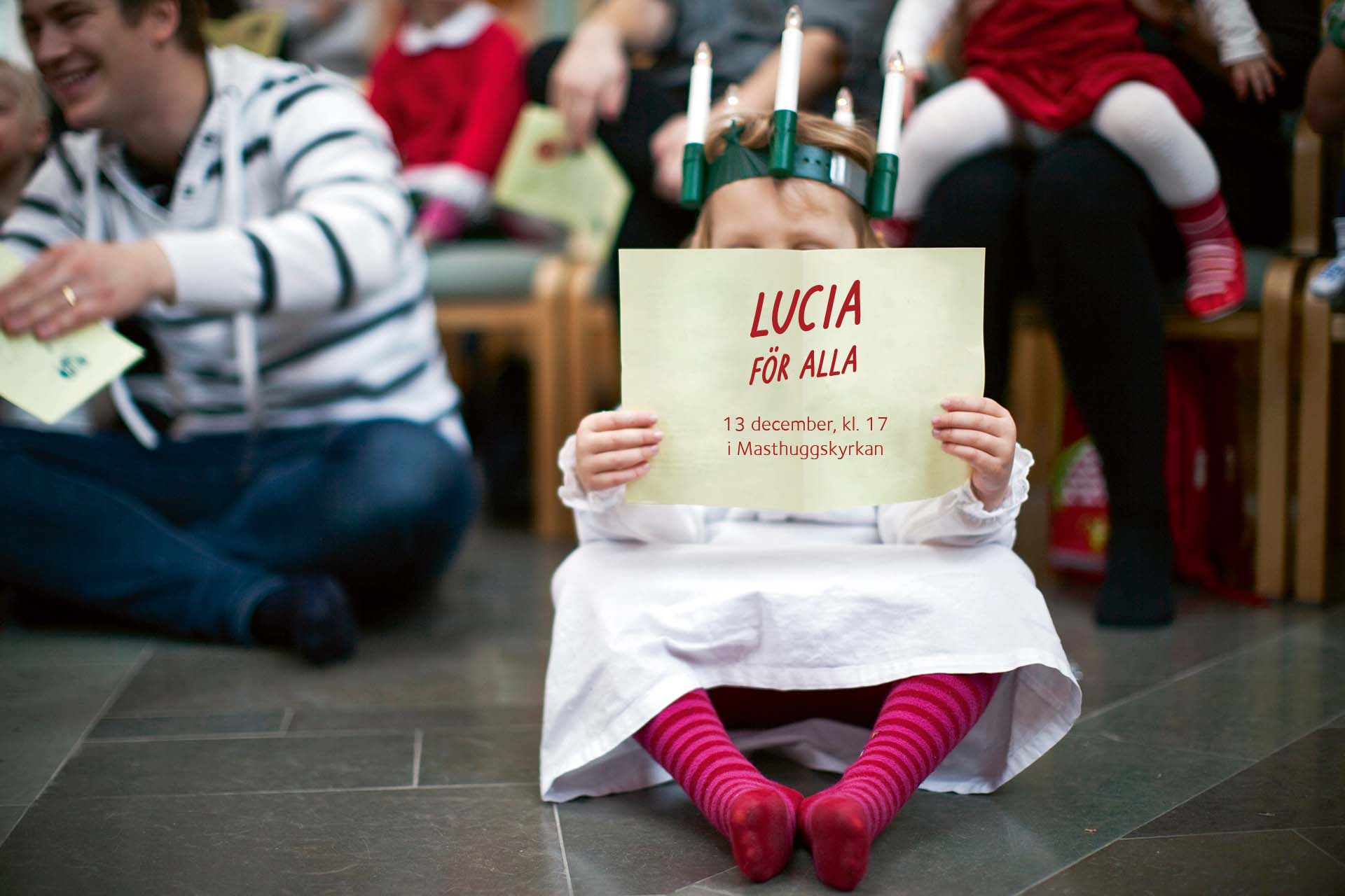 En liten lucia sitter på golvet och läser i ett papper. På pappret står det Lucia för alla.