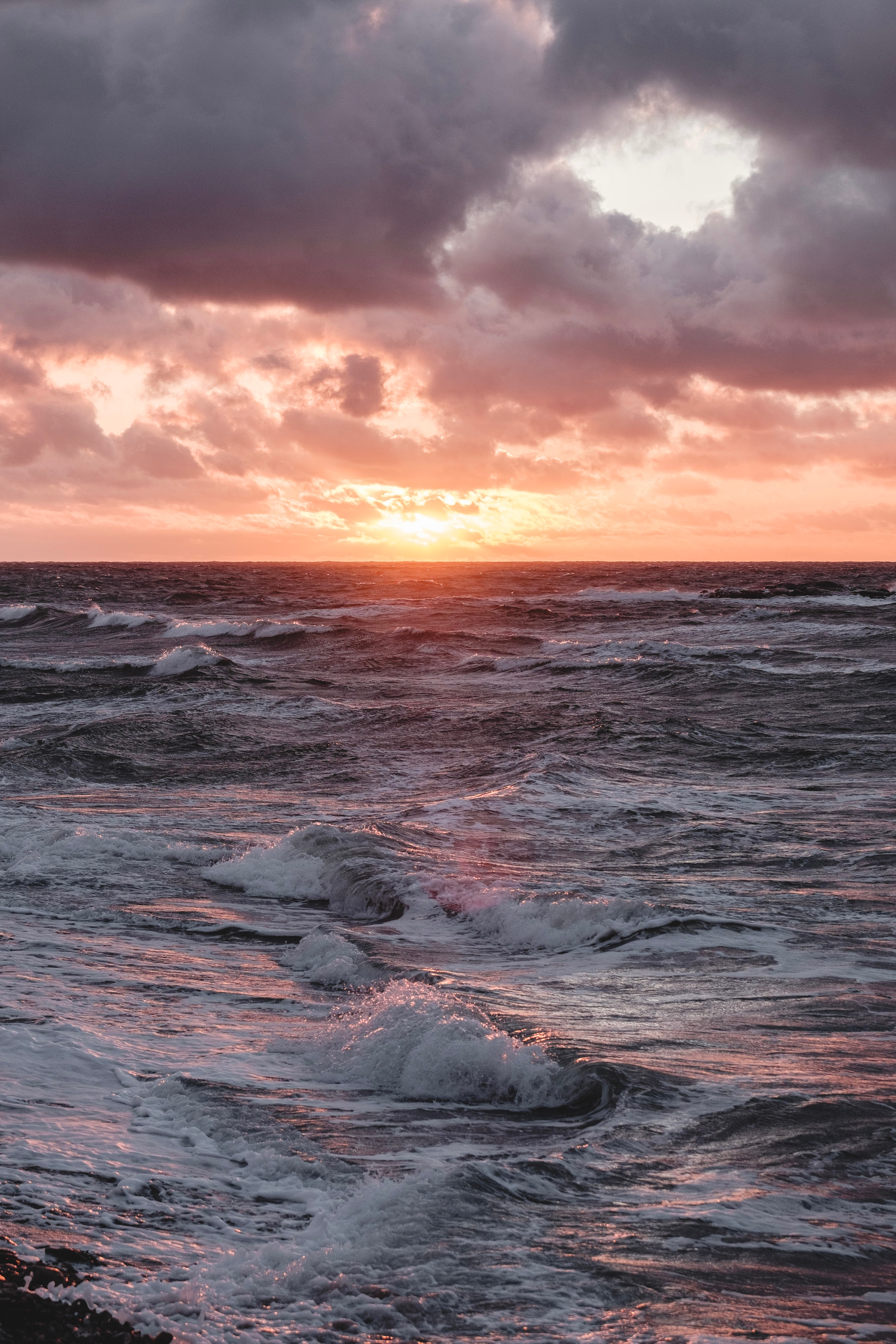 Skummiga vågor på havet. Orange himmel där solen anas bakom molnen.