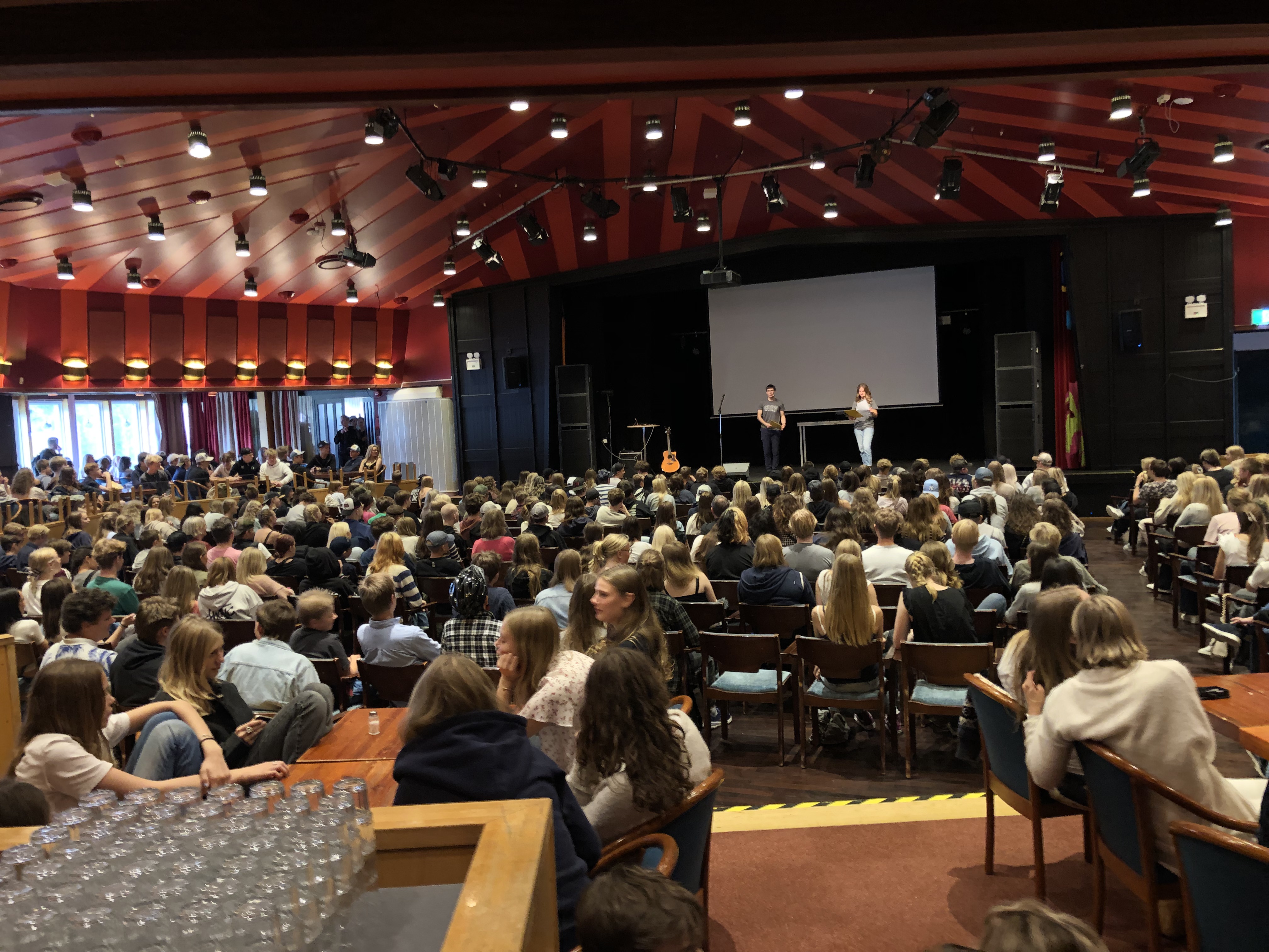 Grupp med ungdomar som lyssnar på två personer som står på scen.