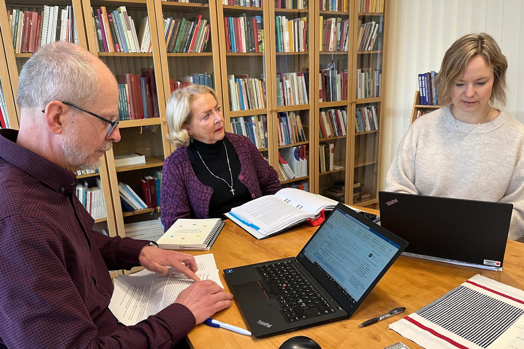 En man och två kvinnor sitter runt ett bord och arbetar med två laptops, papper och pärm.