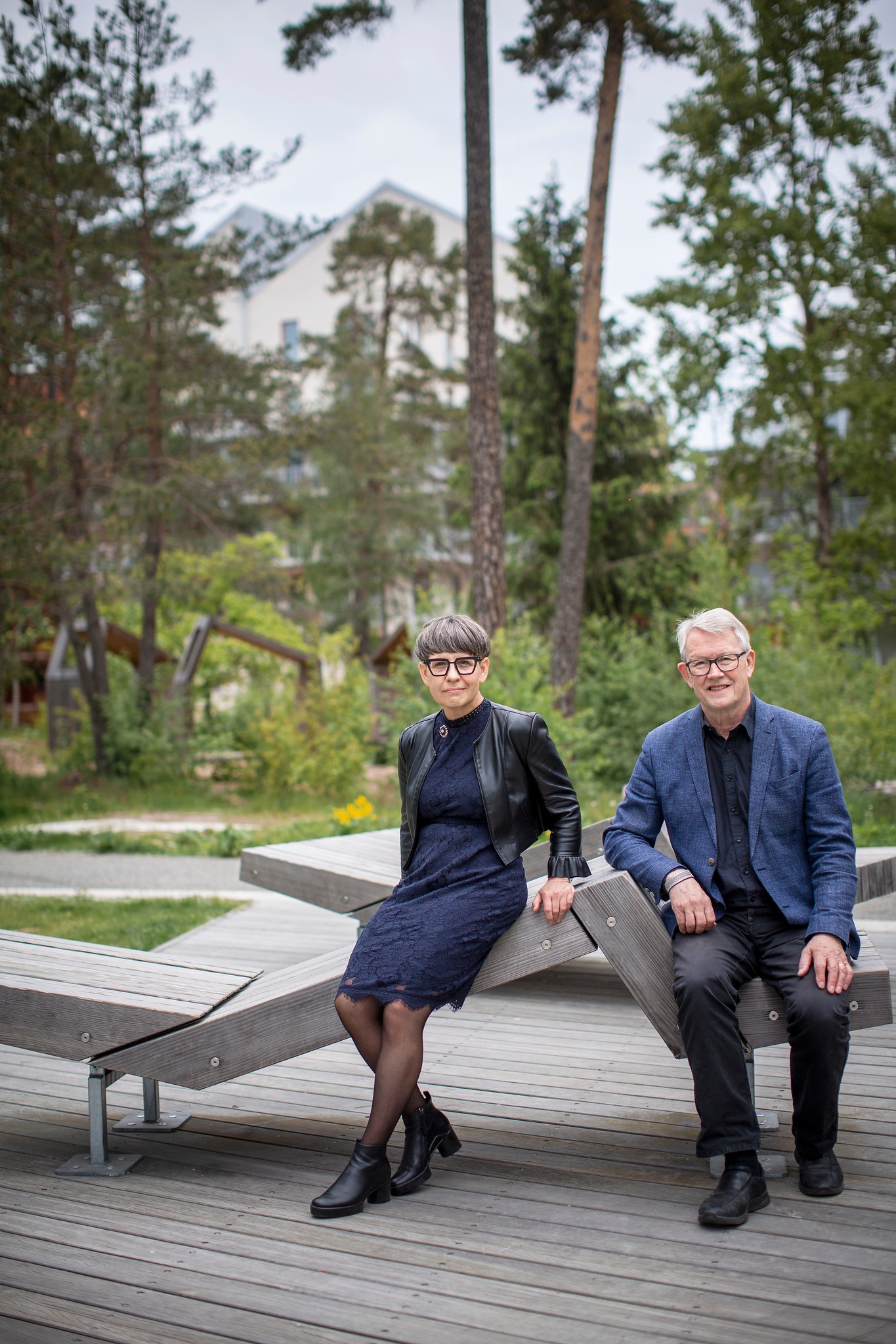 En kvinna och en man, båda uppklädda i klänning respektive skjorta och kavaj, sitter på en träbänk i en skogspark.