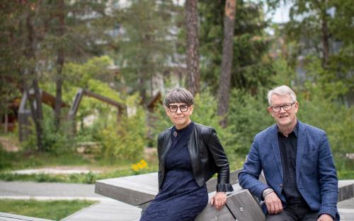 En kvinna och en man, båda uppklädda i klänning respektive skjorta och kavaj, sitter på en träbänk i en skogspark.