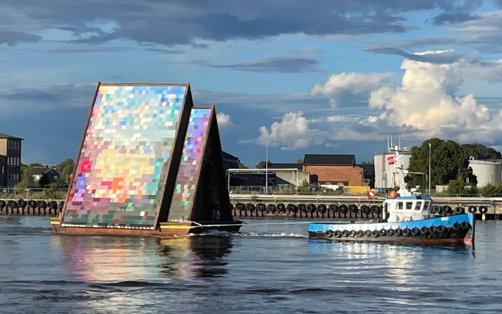 En lotsbåt bogserar pråmkatedralen förbi en kaj med hus.