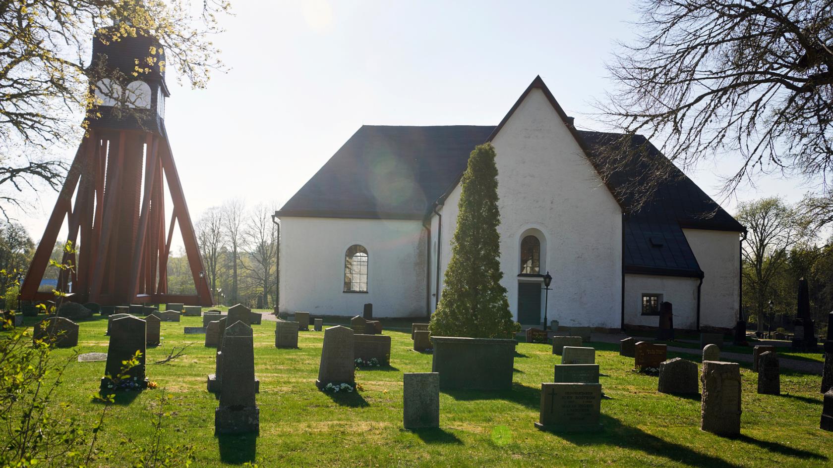 Solen skiner ner på ett klocktorn i rött och en vit ganska kompakt kyrka. I förgrunden ser vi kyrkogården med gravstenar och grönområden.